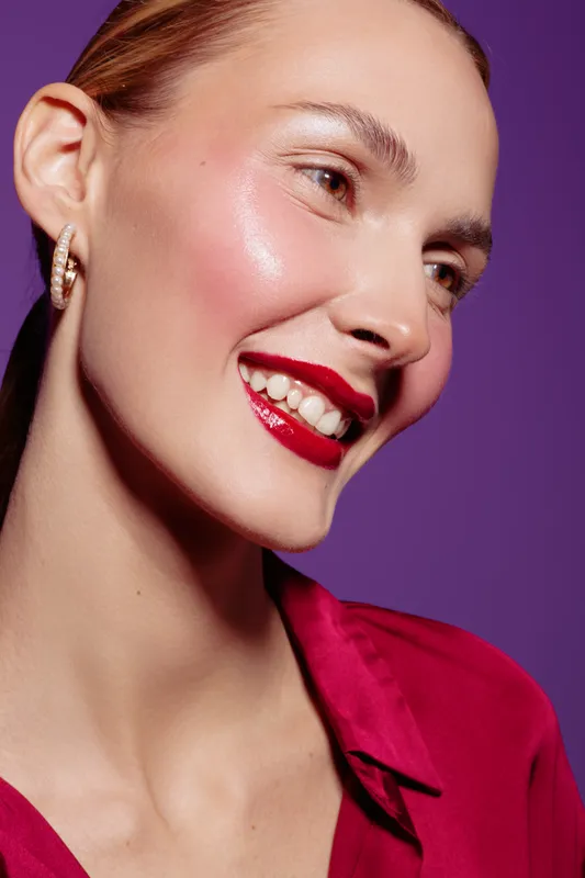 Smiling woman with red lipstick and glowing skin wears red shirt against purple background. Close-up portrait with natural makeup and joyful expression. Concept of beauty and fashion.