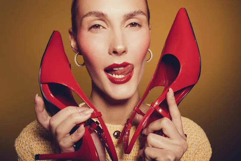 Close-up of a woman holding red high heels near her face with playful expression and tongue out. Fashion concept with bright colors and stylish accessories on yellow background.