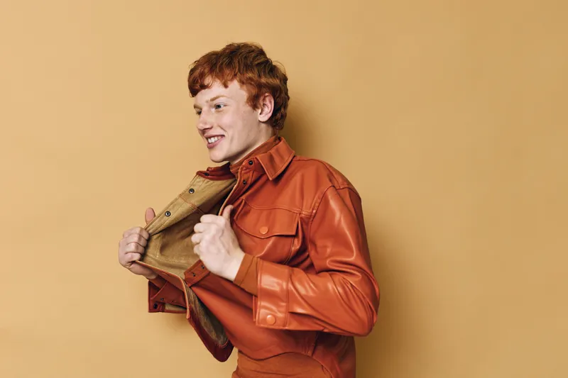 Young man with red hair wearing orange leather jacket, smiling and posing against beige background with happy expression and casual style in studio shot.