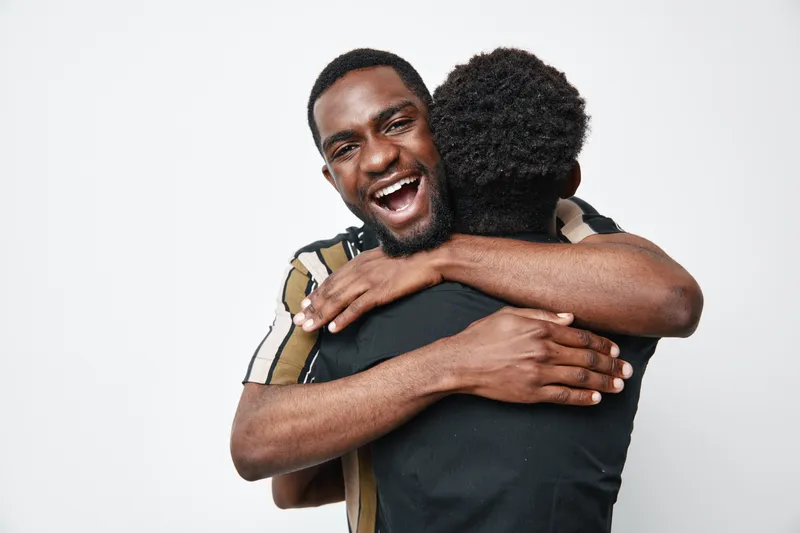 Two young black men hugging each other with happy expressions on white background. African ethnicity, dark skin, friendship and emotional connection between friends or family.