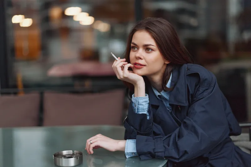 Young woman in a denim jacket sits at a cafe table smoking a cigarette and gazing thoughtfully, with muted urban background and soft evening light around her glow.