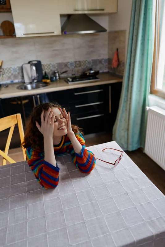Woman indoors at a kitchen table, distressed expression, hands on temples, colorful striped sweater, natural light, home lifestyle moment, cluttered counter, calm colors