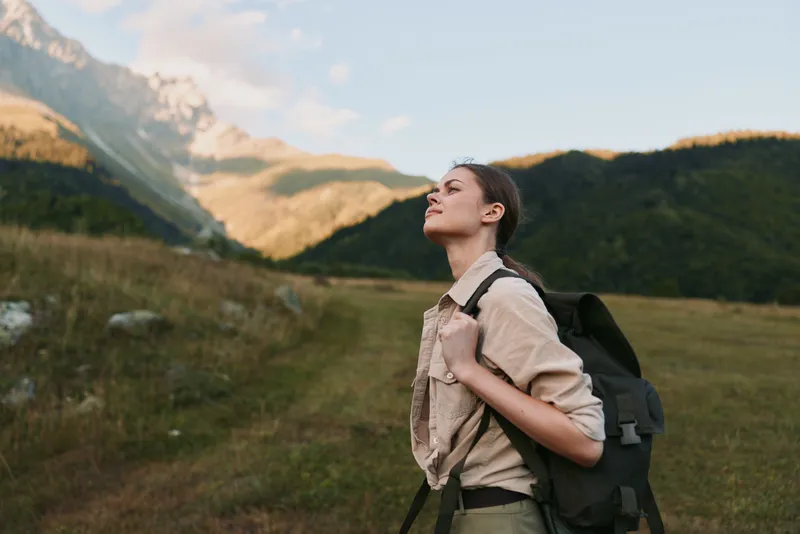 A woman hiker with a backpack gazes up at towering mountains, savoring the wild outdoors in a vast landscape, a moment of journey, exploration, and freedom.