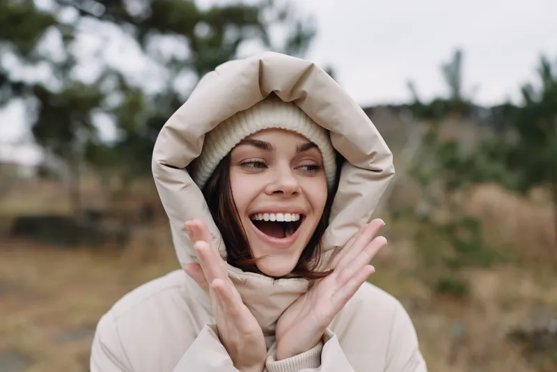 Smiling woman in a cozy hoodie outdoors in a chilly landscape, joyful expression with hands near face, winter wardrobe amid natural scenery.