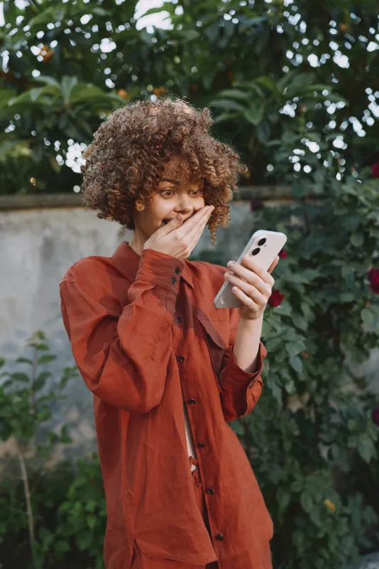 Woman lifestyle outdoors uses smartphone, expressing surprise during a casual fashion portrait in a garden, smiling candidly under natural sunlight, highlighting curly hair and rust shirt