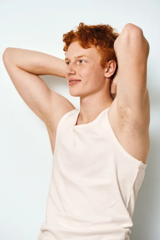 Young man with red hair wearing white sleeveless shirt stretching arms behind head looking away on white background in casual clothing with relaxed expression and natural pose.