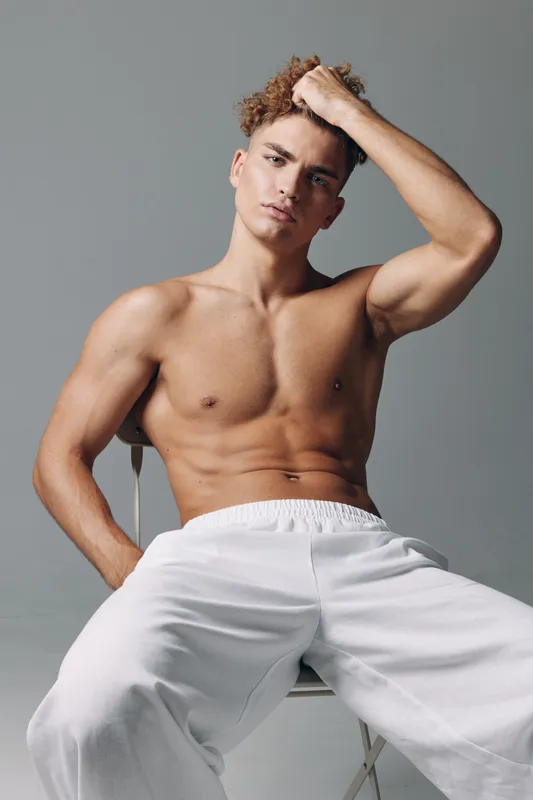 Young muscular man with curly hair sitting shirtless on chair wearing white pants, posing with hand on head against gray background in studio lighting.