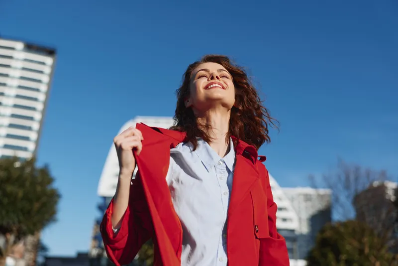 A woman in a red coat stands outdoors, smiling as she looks up at a clear blue sky. A bright urban backdrop with modern buildings adds energy and optimism.