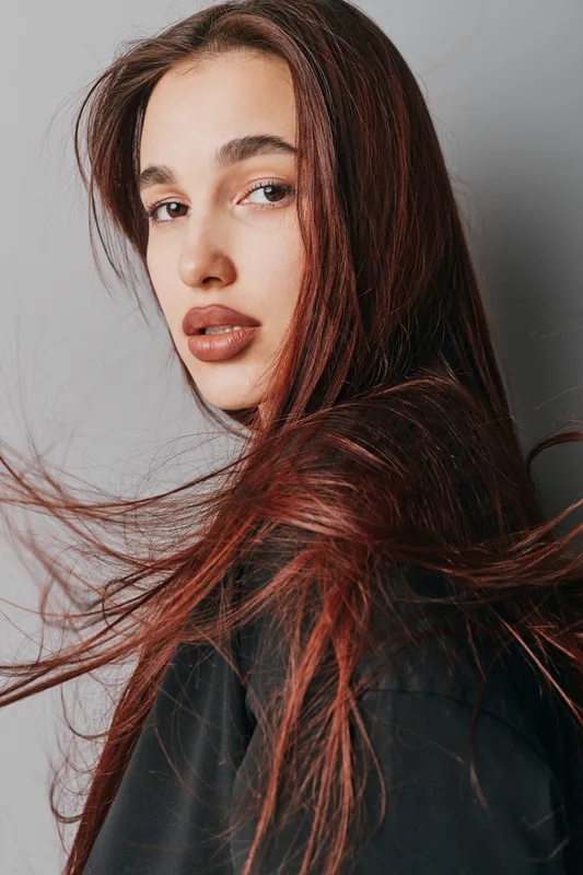 Woman with long red hair and natural makeup posing against gray background. Portrait of young female with loose hair strands and serious expression wearing dark clothing.