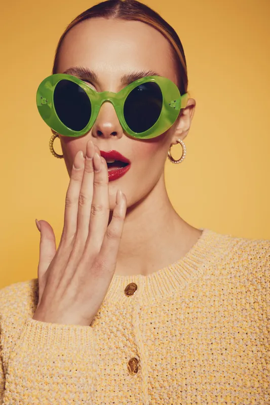 Stylish woman with green round sunglasses and red lipstick covers mouth with hand. She wears yellow knitted sweater and gold hoop earrings on yellow background. Concept of fashion and surprise.