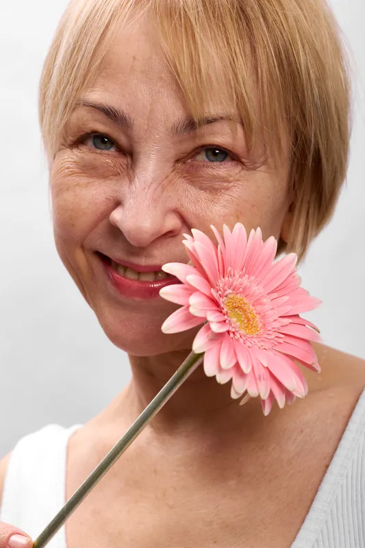 Smiling blond woman closeup portrait holding pink gerbera flower near cheek gentle expression, light gray background, spring mood, soft focus, hair visible