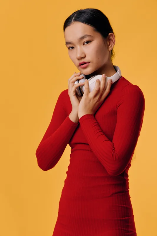 Young woman in red dress with headphones around her neck posing against yellow background. Portrait of girl with dark hair wearing casual outfit in studio setting with confident expression.