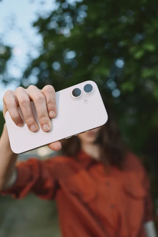 A woman outdoors takes a selfie with a white smartphone, capturing casual everyday life. The image highlights modern female lifestyle, self expression, and connection in a sunny outdoor setting