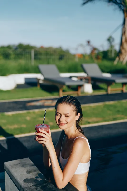 Woman in a white bikini sits by a pool sipping a vibrant drink, sunny outdoor scene, poolside leisure, self care moment, relaxed smile, warm weather, wellness vibe, casual summer lifestyle.