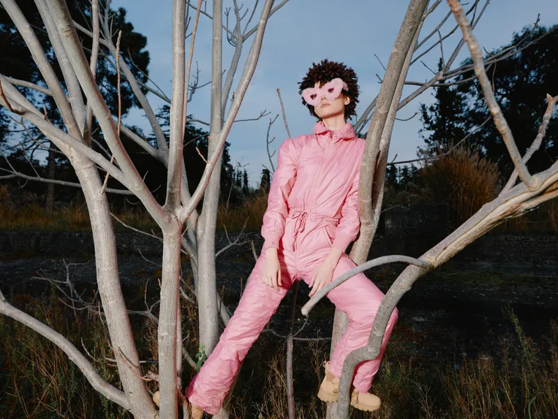 Doll in pink jumpsuit clings to barren tree branches in an outdoor setting, wearing a dark mask and eerie expression, creating a surreal, haunting art installation vibe.