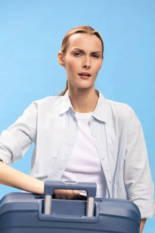 Confused woman with light hair and white shirt holds blue suitcase handle against blue background. Concept for travel uncertainty, doubt, and planning challenges.