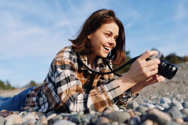 Camera, photography, outdoors, woman, lying, pebbles, beach, hobby, lens, smiling, candid moment under sunny sky with camera in hand, capturing a joyful outdoor scene during a relaxed photoshoot on