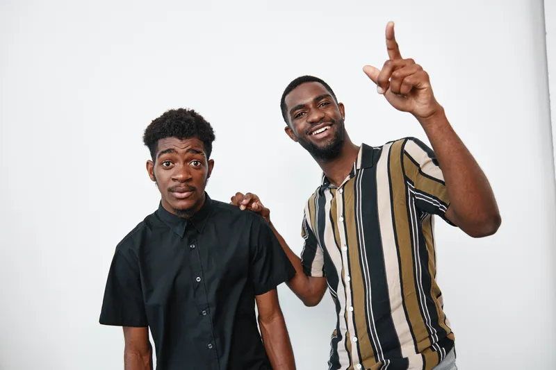 Two african men with dark skin standing together on white background one smiling and pointing up while the other looks at camera wearing casual shirts indoors.