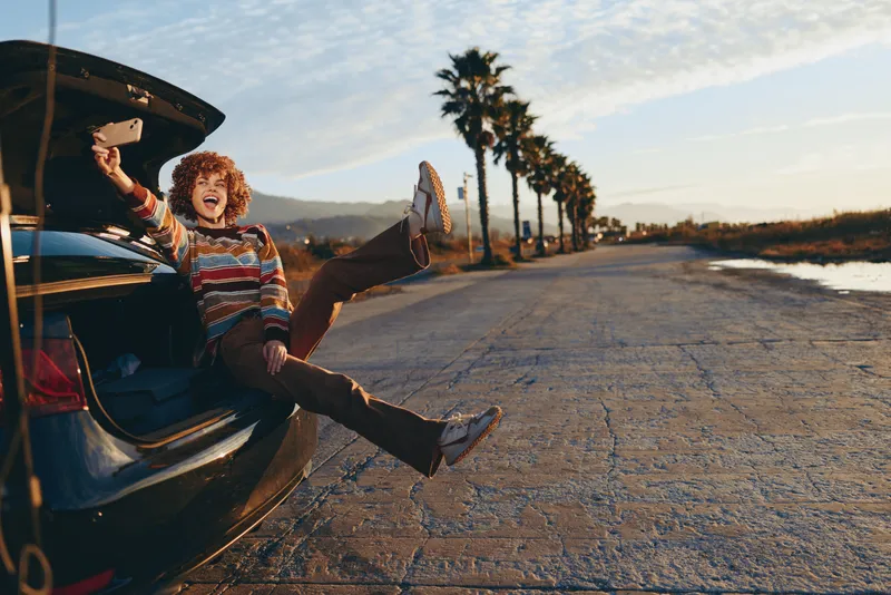 Playful woman lifestyle beside open car trunk on a quiet road at sunset, joyful expression with legs up on the trunk, relaxed posture, near palm lined avenue in countryside for road trip