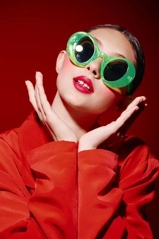 Fashion model posing with oversized green sunglasses and glossy red lips, wearing a bright red coat on a red background. Creative beauty concept focused on unique style and color contrast.