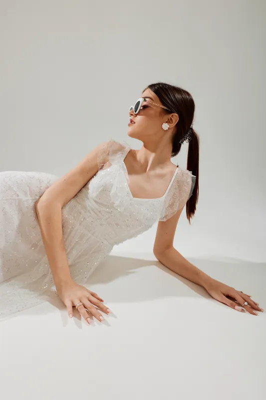 Fashionable woman in a white dress poses reclining on a white surface wearing stylish sunglasses and earrings. Concept of beauty, elegance, fashion, and modern style in studio lighting.