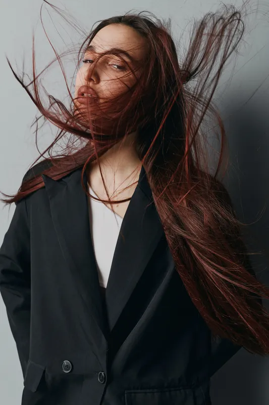 Woman with long hair blowing in wind wearing black blazer and white top posing against gray background in studio for fashion portrait and style concept.