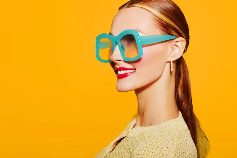 Stylish woman with long hair wearing oversized blue sunglasses and red lipstick smiles confidently against bright yellow background. Concept of fashion, beauty, and summer style.