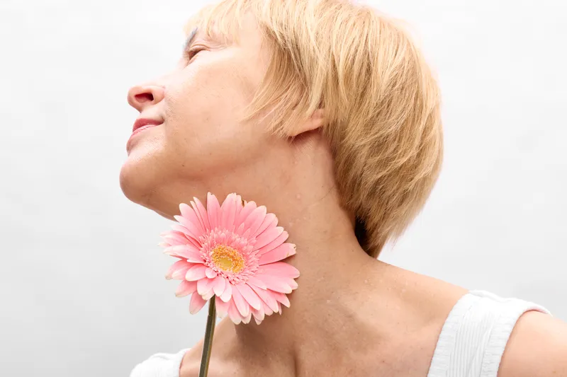 Woman portrait in side profile, blonde hair, eyes closed, holding a pink gerbera flower near cheek, calm spring mood, soft white background, close up photography, copy space