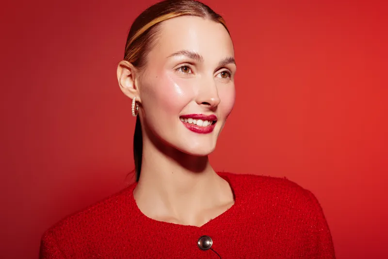 Smiling woman with slicked back hair wears red sweater and earrings against red background. Studio portrait with bright lighting and glossy skin. Concept of fashion and beauty.