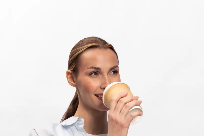 Young woman with tied hair drinking coffee from a paper cup isolated on white background. Concept of morning routine, refreshment, and casual lifestyle with natural expression.