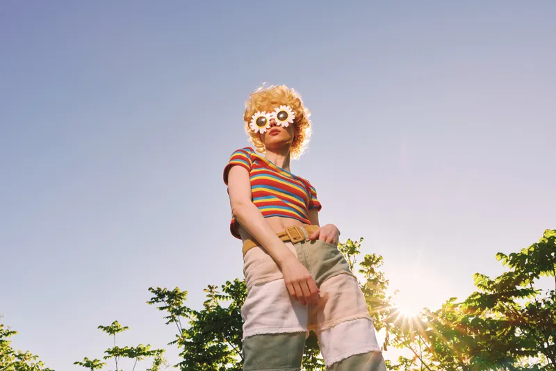 Smiling woman with curly hair wearing a colorful striped top and flower sunglasses outdoors. Vintage vibe with warm retro film color and bright sunlight, capturing carefree lifestyle and playful