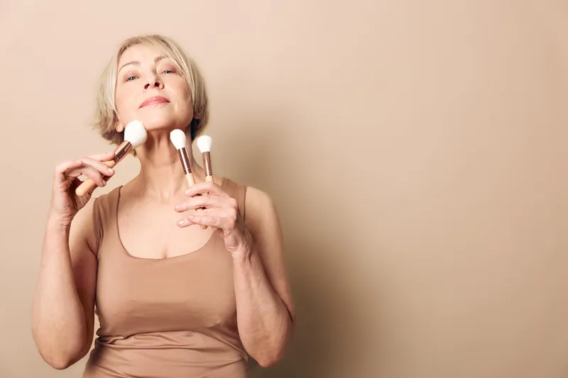 Confident mature woman with short hair applies makeup brushes on face against neutral background. Concept of beauty routine, skincare, self-care, and ageless elegance in natural light.