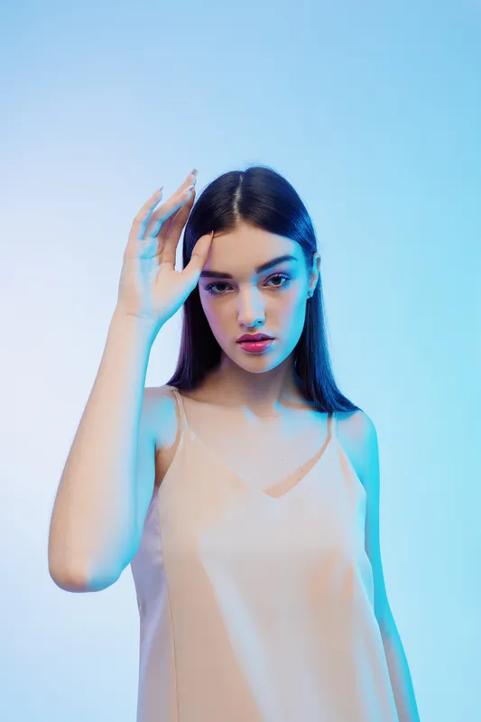 Young woman beauty fashion model poses with hand near face under blue studio lighting. Concept for style, elegance, and modern portrait photography.