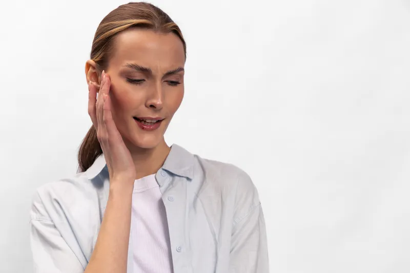 Young woman with closed eyes touches her temple with hand, expressing headache or stress. White background emphasizes emotion of discomfort and tension in casual clothing.