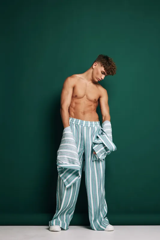 Young shirtless man wearing striped pajama pants and holding matching pajama top standing against dark green background with casual relaxed pose and bare feet.