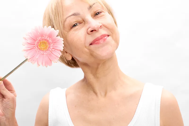 Senior woman holding pink gerbera flower near face, smiling, closeup on white studio background, beauty and skincare concept, calm expression, soft makeup, fresh
