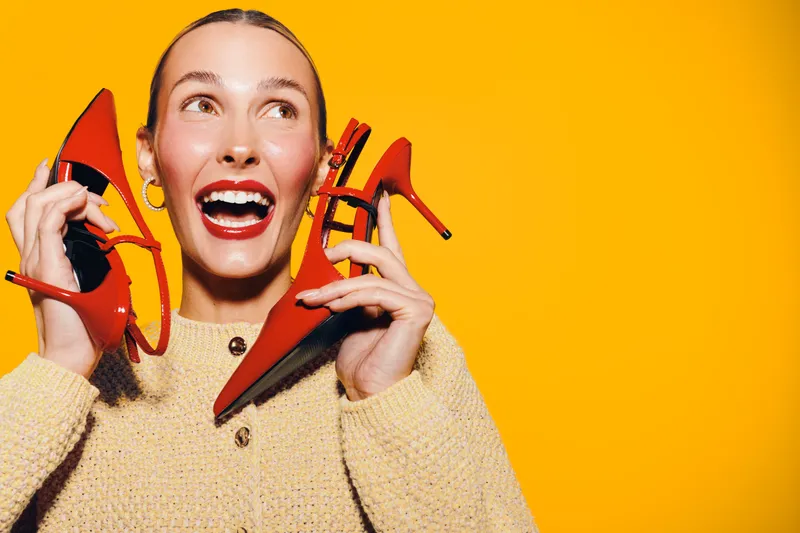Happy woman with red high heels holds shoes near face, smiling broadly on bright yellow background. Concept of fashion, style, shopping, and joyful emotions in studio lighting.