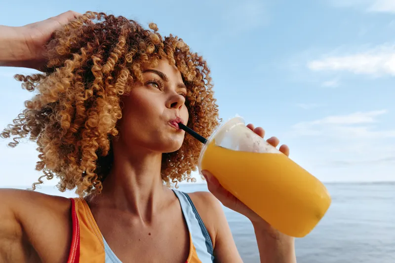 Young woman with curly hair drinks orange juice on a sunlit beach, wearing a colorful tank top, enjoying a casual, carefree moment by the sea in a light breeze.