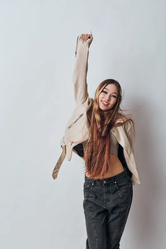 Young woman with long hair wearing jacket and jeans smiling and raising hand in studio with white background casual fashion and happiness expression lifestyle portrait.
