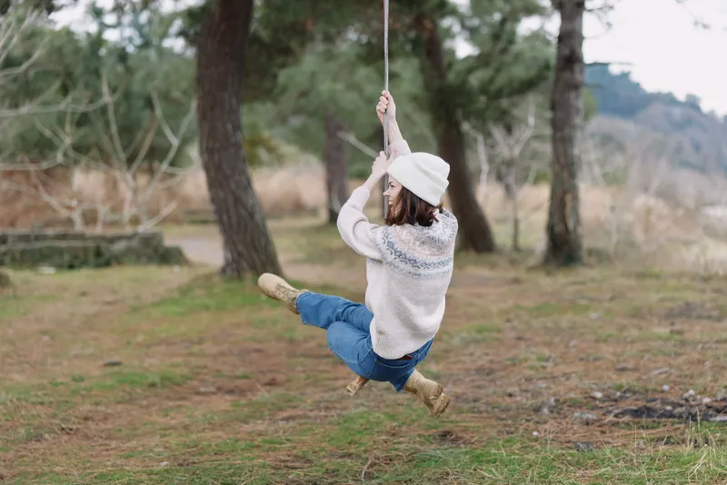 Girl swinging on a rope swing in a park, wearing a knit sweater, hat and boots, mid motion with trees and grass in the background, capturing carefree outdoor play in nature