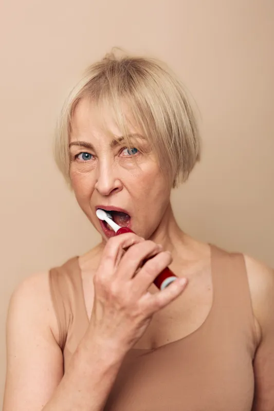 Middle aged woman with short blonde hair brushing teeth with electric toothbrush in bathroom. Close up portrait on beige background. Concept of dental care, hygiene, and healthy lifestyle.