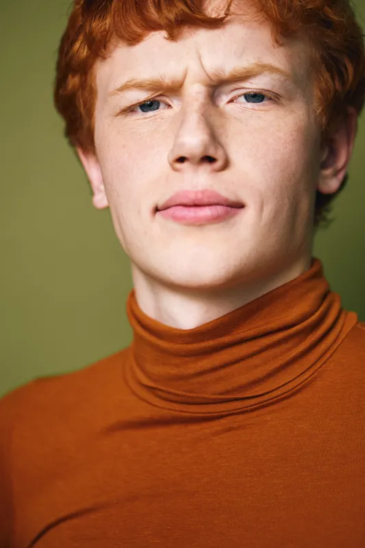 Closeup portrait of young redhead man with freckles and curly hair wearing brown turtleneck looking at camera with serious expression and furrowed brow against green background.