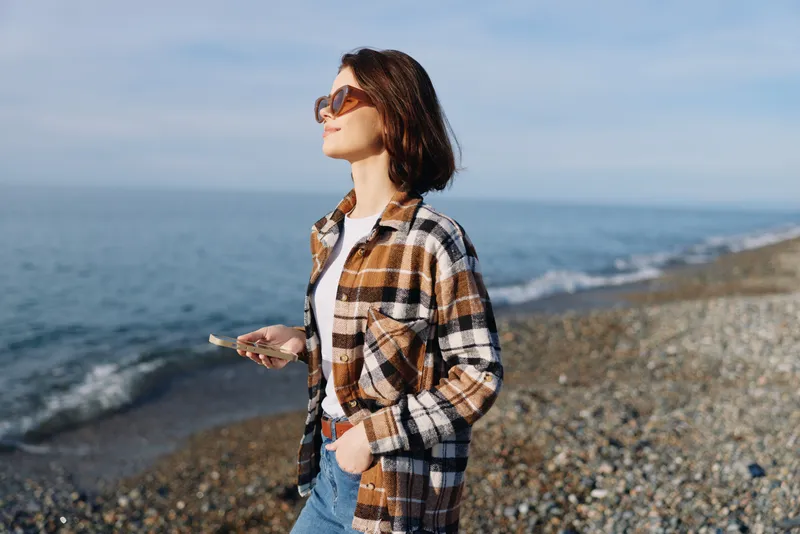 Woman on a pebbled beach wearing a plaid shirt and sunglasses, holding a phone, gazing toward the sea in a casual outdoor moment.