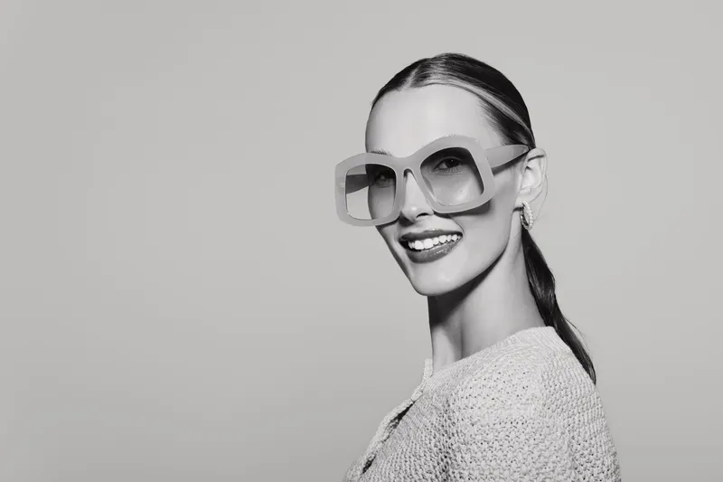 Smiling white woman with pale skin and slicked hair wearing large sunglasses and a knitted sweater. Studio portrait in black and white with confident mood.