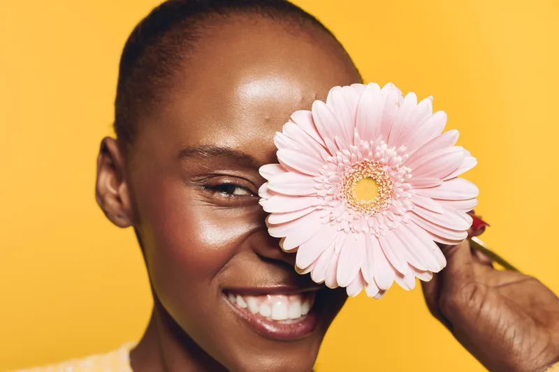 Close-up of a smiling dark skinned African woman holding a pink flower over one eye. Yellow background highlights natural beauty, happiness, and floral concept in studio lighting.