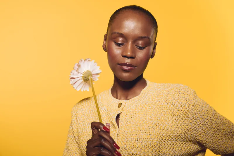 Portrait of a calm African woman with dark skin holding a white flower against yellow background. Concept for natural beauty, serenity, and floral wellness in studio lighting.