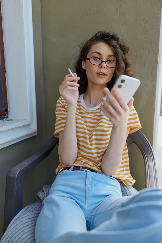 Young woman with short wavy hair sits on a porch, holding a cigarette in one hand and a smartphone in the other, wearing a striped yellow tee and light blue jeans, conveying casual relaxed mood.