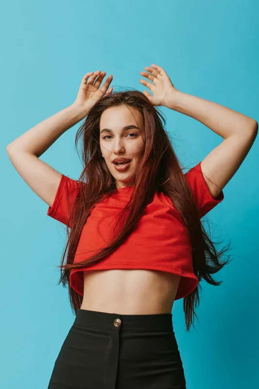 Young woman with long hair wearing red crop top and black pants posing with hands raised against blue background. Casual style, confident expression, studio portrait with bright colors.