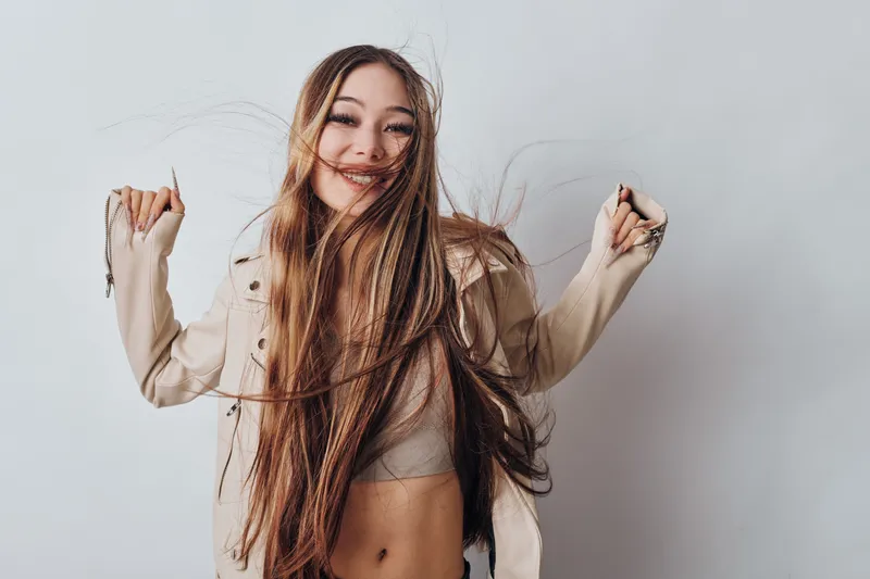 Young woman with long braided hair smiling and dancing in casual jacket on white background. Portrait of happy female with carefree expression and natural beauty indoors.