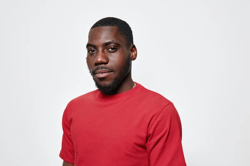 Portrait of confident black man with dark skin wearing red t shirt standing against white background looking at camera with slight smile and relaxed posture.